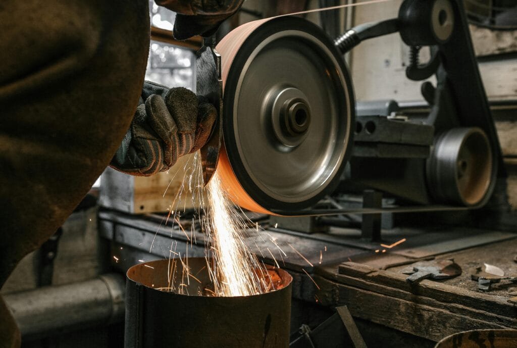 A close-up of a metalworker grinding a blade, creating sparks in a workshop setting.